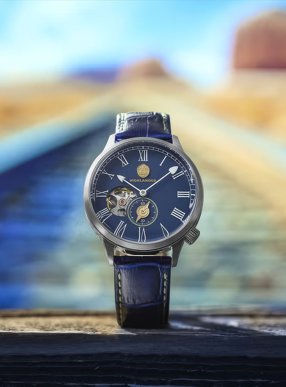 A luxury wristwatch with a dark blue dial, Roman numeral hour markers, a skeletonized side window, and a matching blue leather strap, standing upright on a dark wooden surface with a blurred desert landscape background.