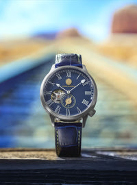 A luxury wristwatch with a dark blue dial, Roman numeral hour markers, a skeletonized side window, and a matching blue leather strap, standing upright on a dark wooden surface with a blurred desert landscape background.