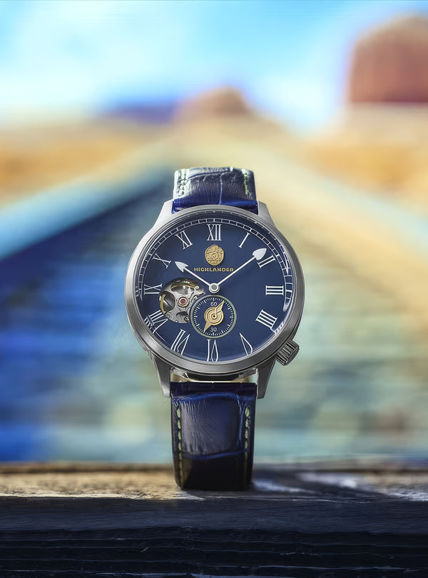 A luxury wristwatch with a dark blue dial, Roman numeral hour markers, a skeletonized side window, and a matching blue leather strap, standing upright on a dark wooden surface with a blurred desert landscape background.