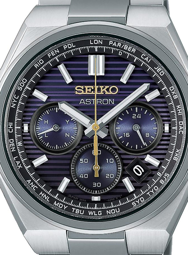A close-up of a Seiko Astron wristwatch with a brushed silver-tone metal case and bracelet, featuring a dark blue and purple striped dial with white, silver, and gold details.